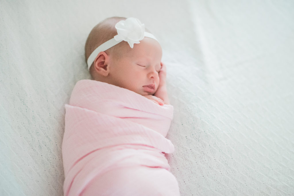 newborn baby girl swaddled in pink aden + anais blanket and white bow