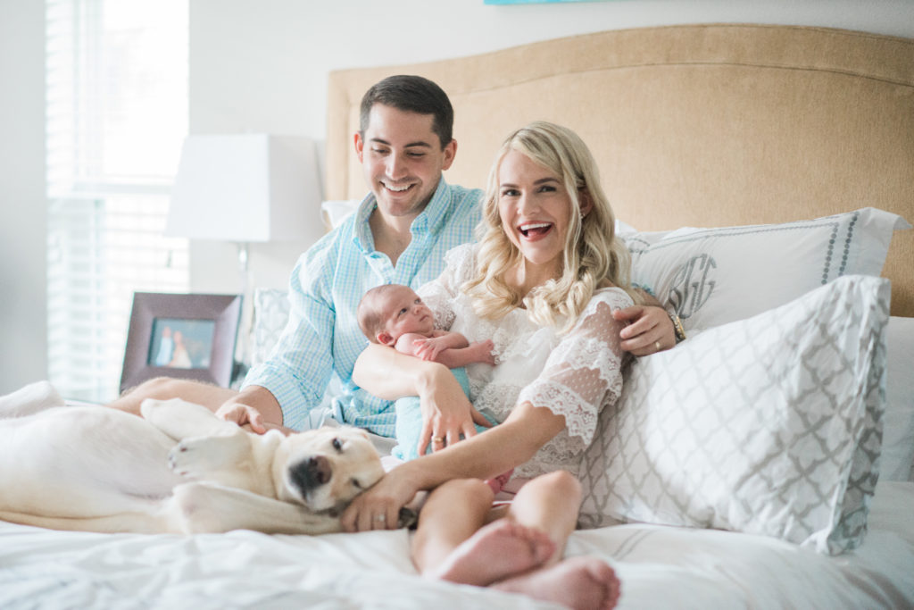 mom in white lace blouse holding newborn baby next to husband in blue button down with yellow lab all on bed