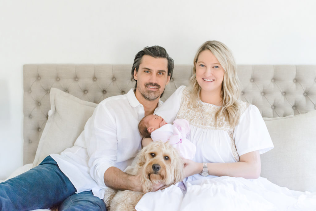 mom in white dress, dad in white button down, baby girl in Beaufort bonnet swaddle & dog on neutral bed